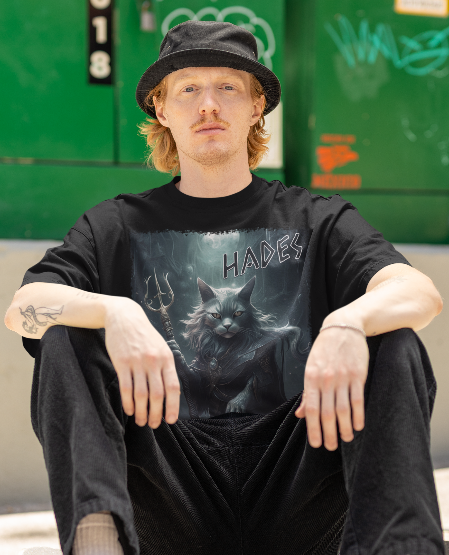 Person wearing a black t-shirt with a graphic design, sitting on a skateboard with a graffiti-covered wall in the background.